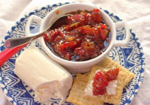 A bowl of tomato and pepper jam with cheese and crackers on a blue and white patterned plate.