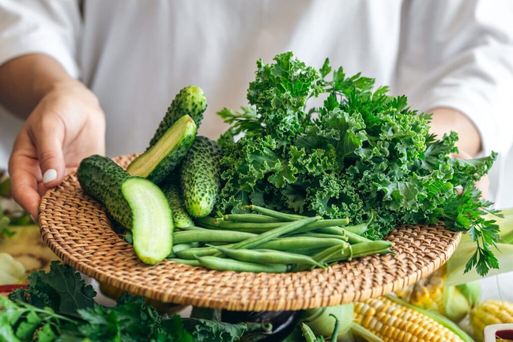 A woman holds a plate with cucumbers, kale, parsley and green beans